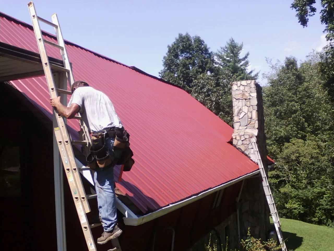 Roofer on a ladder beside a red metal roof and stone chimney — Soffit & Fascia Repair by Roofing Brothers in Dorothy